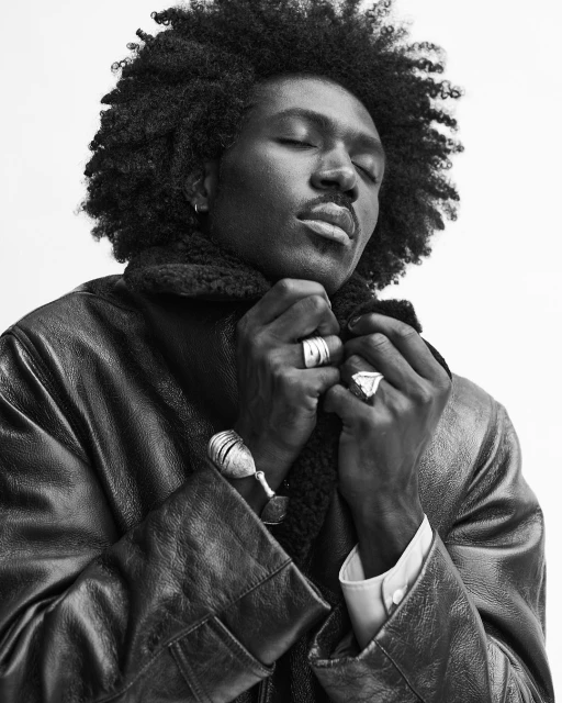 Black and white studio portrait of a Black man with curly hair wearing a jacket, eyes closed, hands at his collar, against white.