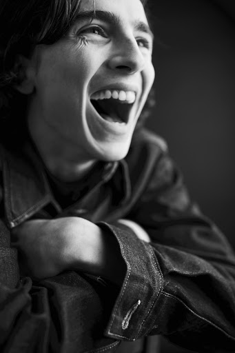 Black and white portrait of Timothee Chalamet laughing heartily with head tilted back and arms crossed on a dark background