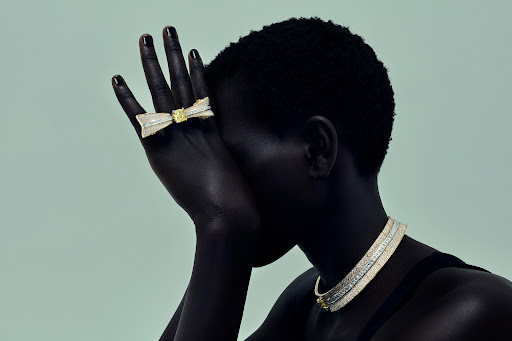 Black woman wearing an elaborate diamond bow ring across two fingers and a matching diamond necklace against a pale green background