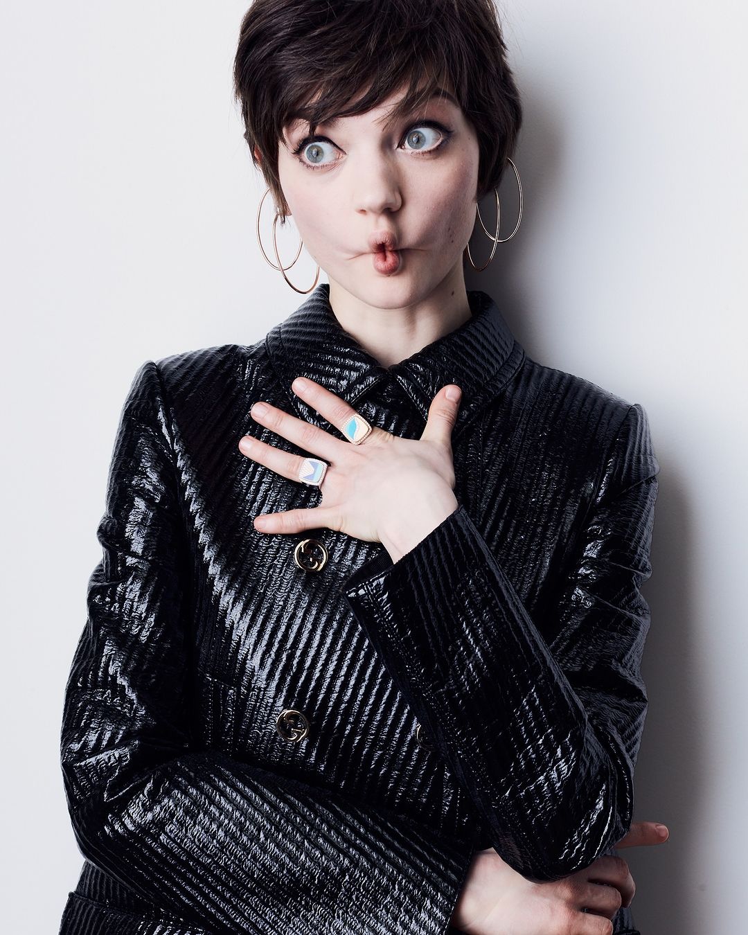 A young woman with short dark hair, large hoop earrings, and a shiny black jacket strikes a funny pose against a white background.