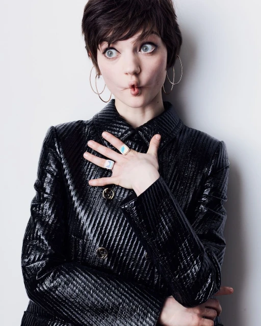 A young woman with short dark hair, large hoop earrings, and a shiny black jacket strikes a funny pose against a white background.