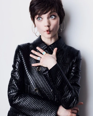 A young woman with short dark hair, large hoop earrings, and a shiny black jacket strikes a funny pose against a white background.