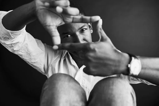 Monochrome portrait of an African American man framing his face with hands, wearing a light shirt and watch against a dark background.