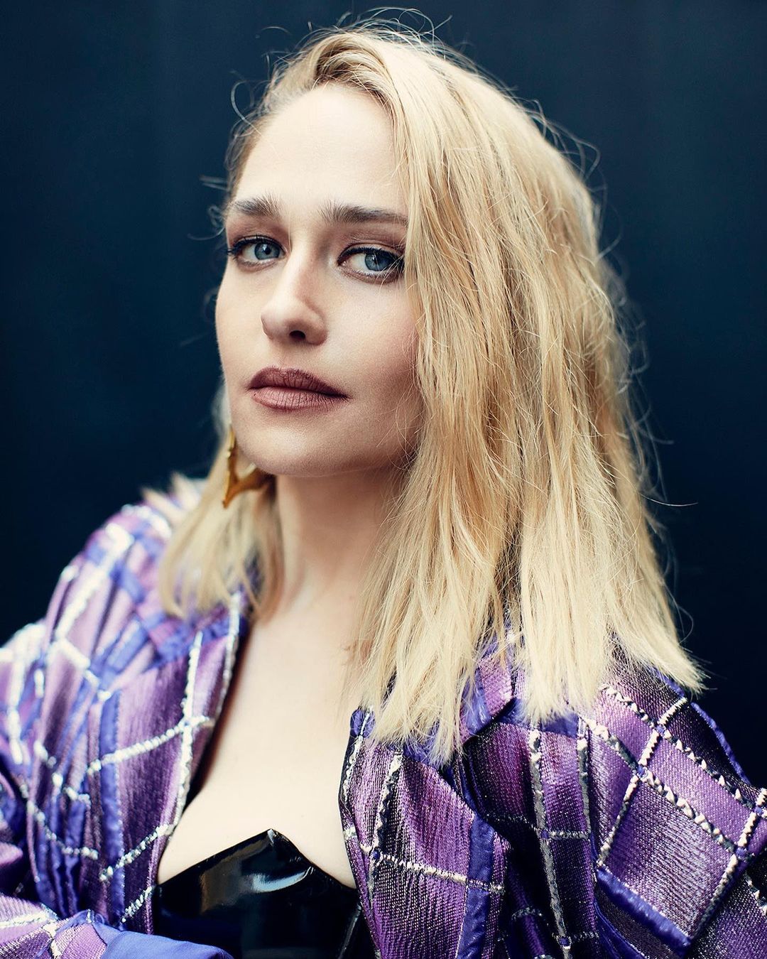 Close-up portrait of an adult female with blonde hair and blue eyes, wearing a purple patterned jacket against a dark background.