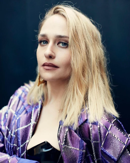 Close-up portrait of an adult female with blonde hair and blue eyes, wearing a purple patterned jacket against a dark background.