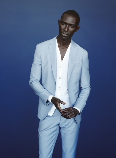 Stylish African male fashion model poses in a light blue suit with a white vest against a complementary blue background in a studio shot.
