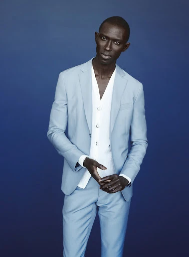 Stylish African male fashion model poses in a light blue suit with a white vest against a complementary blue background in a studio shot.