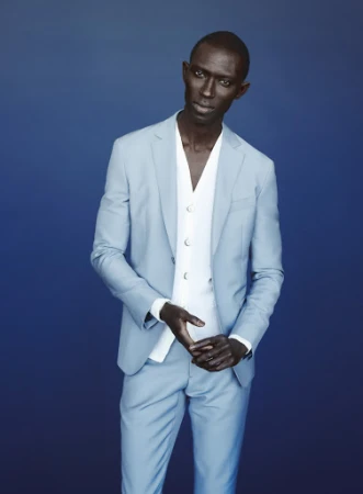 Stylish African male fashion model poses in a light blue suit with a white vest against a complementary blue background in a studio shot.