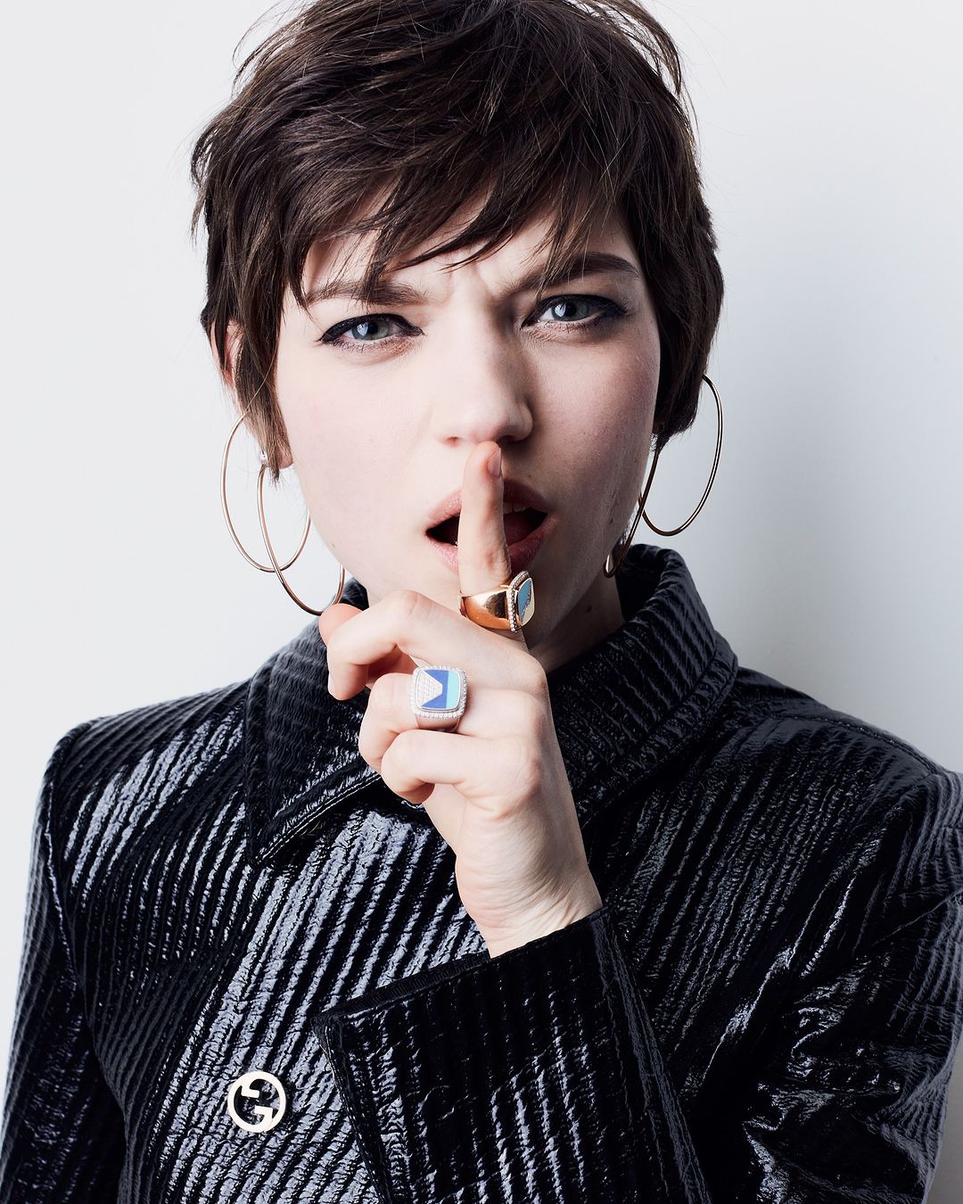 Woman with short brown hair, wearing a shiny black jacket and gold hoop earrings, holds a finger to her lips in a shushing gesture.
