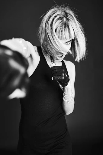 Black and white portrait of a blonde adult woman with boxing gloves throwing a punch in a photo studio with dark background.