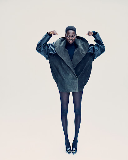 An African American woman in a voluminous dark green coat and black tights and heels jumps against a neutral background in a fashion shot.