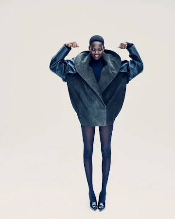 An African American woman in a voluminous dark green coat and black tights and heels jumps against a neutral background in a fashion shot.