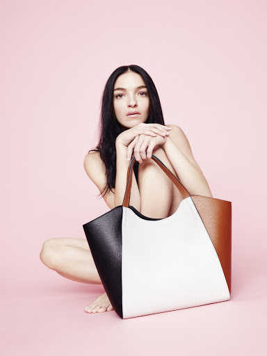 Brunette woman with dark hair squats with a color-blocked tan, white, and black tote bag against a light pink seamless backdrop.