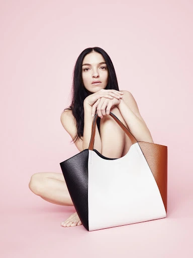 Brunette woman with dark hair squats with a color-blocked tan, white, and black tote bag against a light pink seamless backdrop.