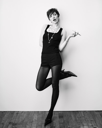 Full-length portrait of a slender young woman in black shorts and a tank top striking a pose against a white wall and wood floor.