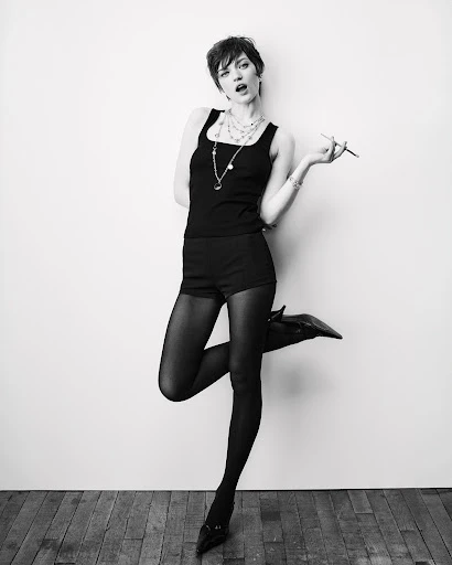 Full-length portrait of a slender young woman in black shorts and a tank top striking a pose against a white wall and wood floor.
