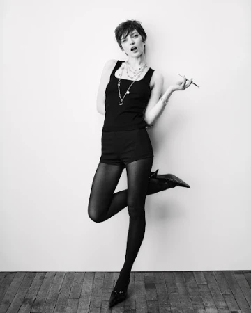Full-length portrait of a slender young woman in black shorts and a tank top striking a pose against a white wall and wood floor.