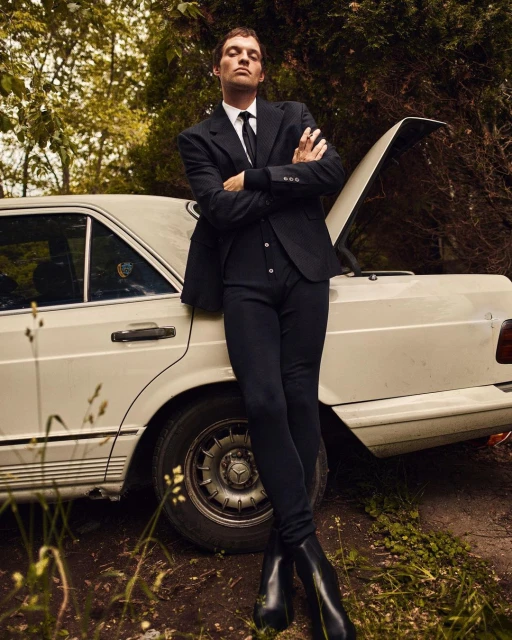 Stylish man in suit poses against vintage car in outdoor setting with green foliage, blending fashion and classic automotive aesthetics.