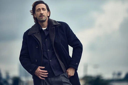 Portrait of an adult man in a pea coat, buttoned shirt, and grey trousers posing against a cloudy sky and blurred cityscape background.