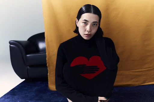 A young adult woman with freckles and slicked-back hair poses in a black turtleneck with a red heart design in a studio setting.