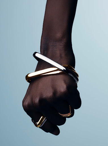 Close-up of a dark-skinned hand wearing metallic gold and silver bangles and rings against a pale blue gradient background.
