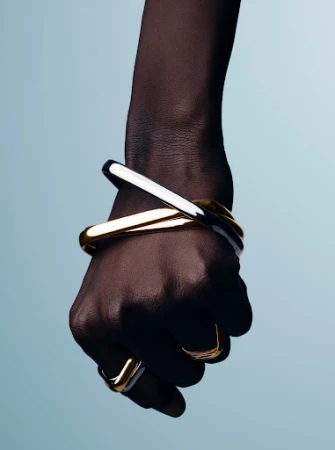 Close-up of a dark-skinned hand wearing metallic gold and silver bangles and rings against a pale blue gradient background.
