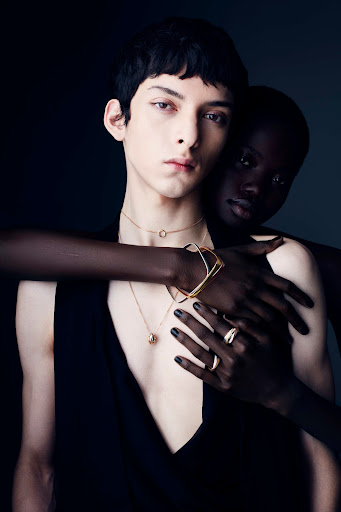 Two people in a close-up studio portrait. One person is fair-skinned with jewelry, and the other is dark-skinned, with arms wrapped around.