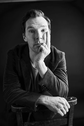 Black and white portrait of Benedict Cumberbatch in jacket, leaning on chair with hand to face, gazing into the camera.