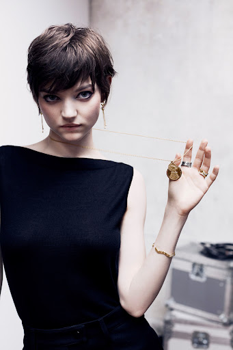 Elegant woman poses with gold jewelry, black top, short hair, and minimal makeup in front of a textured grey background.