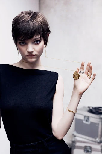 Elegant woman poses with gold jewelry, black top, short hair, and minimal makeup in front of a textured grey background.