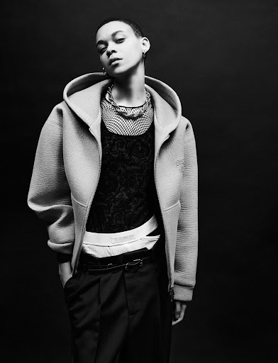 Black and white portrait of a person with short hair wearing a hooded jacket, textured shirt, and dark pants in a studio setting.