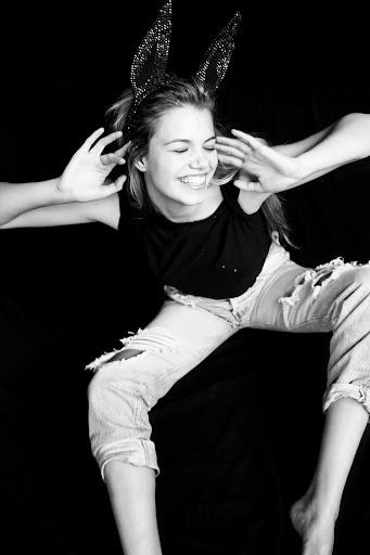 Black and white portrait of a happy woman wearing bunny ears, a black top, and ripped jeans against a dark backdrop.