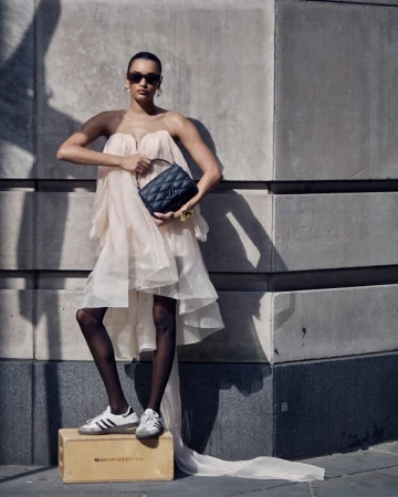 Model in strapless tulle dress and sneakers holding a quilted handbag against a stone building exterior by Billy Kidd