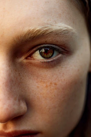 Macro portrait of a woman's face highlighting light amber eyes, blonde eyebrows, and detailed freckles across nose and skin.