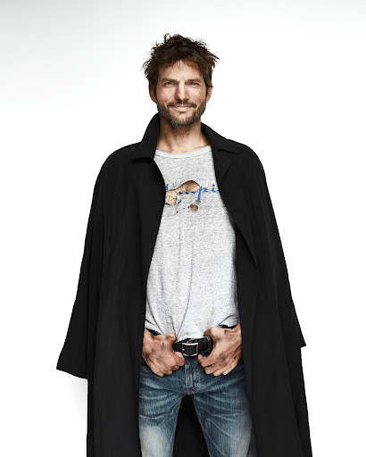 Portrait of a smiling man wearing a black coat, grey t-shirt, and blue jeans against a white background