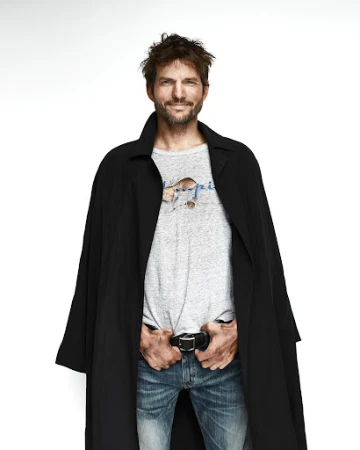 Portrait of a smiling man wearing a black coat, grey t-shirt, and blue jeans against a white background
