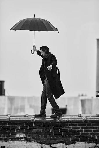 Monochrome image features a man with an umbrella balancing on a brick ledge against a cloudy sky backdrop, creating a dramatic scene.