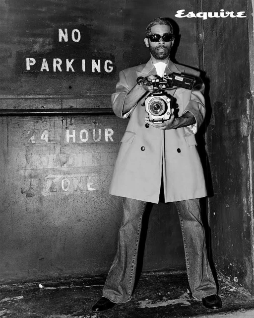 A man holds a video camera in front of a textured door with a 'No Parking' sign, black and white, fashion editorial with magazine logo.