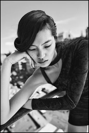 Portrait of a contemplative Asian woman in an elegant black dress, resting her arm on a railing with a blurred city background.