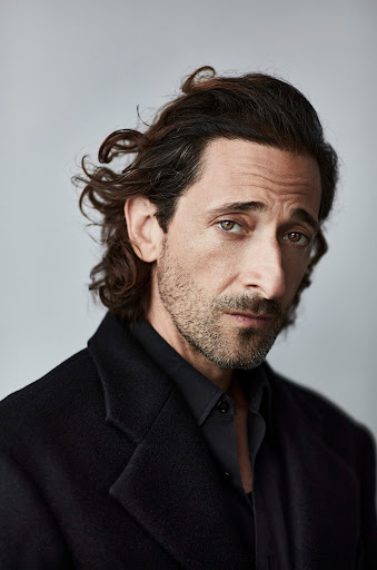 Portrait of actor Adrien Brody in black shirt and coat, looking directly at the camera with a serious facial expression.