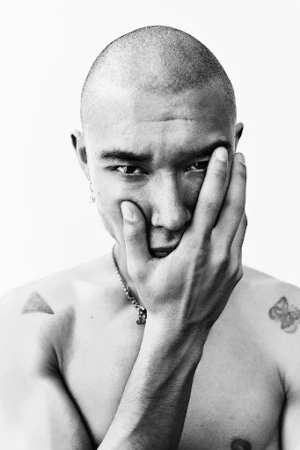 Portrait of a bald young man with tattoos, holding his hand to his face against a white background