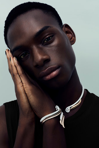 Close-up portrait of dark-skinned male model wearing a modern silver and gold necklace and bracelet against a soft grey background.