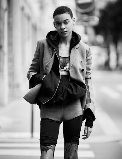 Monochrome portrait of a stylish adult female in a blazer, hoodie, and shorts walking down a city street with pedestrian crossing.