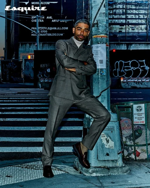 Portrait of an adult man in a grey suit standing on a city sidewalk near a light pole with graffiti, wearing brown leather loafers.