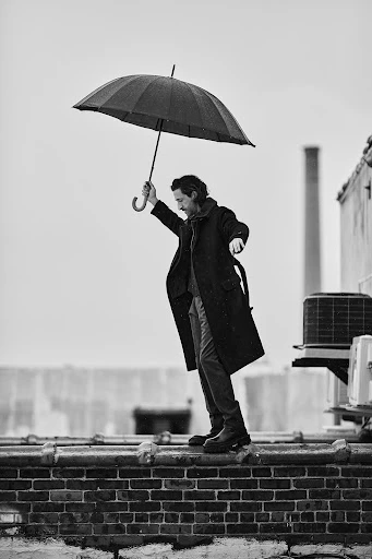 Monochrome shot of an adult man holding an umbrella, wearing a coat and boots, standing on a rooftop ledge against a cloudy sky.