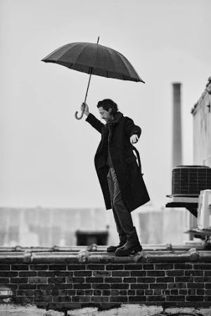 Monochrome shot of an adult man holding an umbrella, wearing a coat and boots, standing on a rooftop ledge against a cloudy sky.