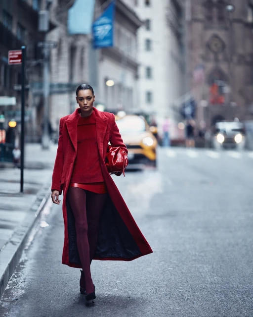 Fashionable woman in red coat and dress walks down a city street with buildings and cars in background on an overcast day.