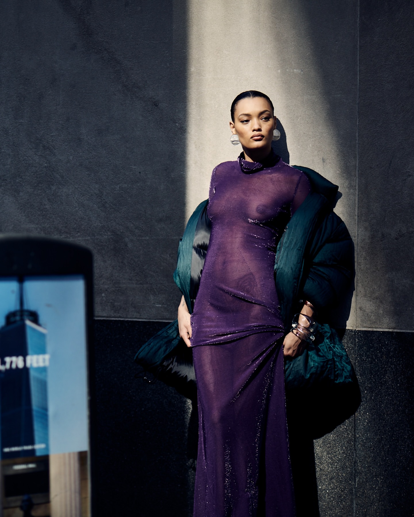Model in a sheer purple dress and dark puffer jacket leaning against a stone wall outdoors, photographed by Billy Kidd