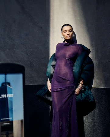 Model in a sheer purple dress and dark puffer jacket leaning against a stone wall outdoors, photographed by Billy Kidd