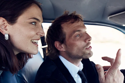 A smiling adult woman and adult man, a couple, ride in a car, shot with shallow depth of field and natural lighting indoors.